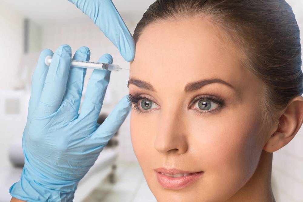 A healthcare professional wearing blue gloves administers a cosmetic injection to a woman&apos;s forehead in a clinical setting. The woman has a smooth complexion and is looking directly at the camera, showcasing her natural beauty. The background is softly blurred, emphasizing the procedure. This image highlights the growing trend of cosmetic treatments aimed at enhancing facial aesthetics.