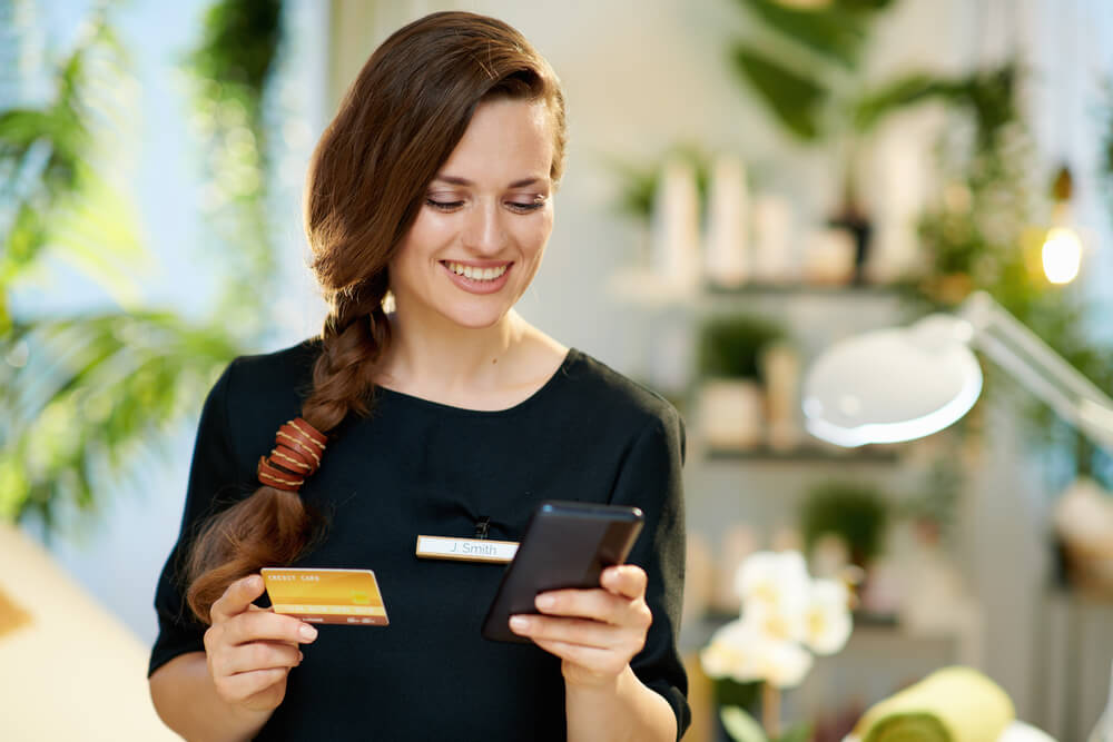 A smiling woman with long brown hair, wearing a black top, holds a credit card in one hand and a smartphone in the other. She appears engaged in a digital transaction or online shopping, surrounded by a bright, plant-filled environment. The image conveys a sense of modern retail and customer service, highlighting the integration of technology in everyday shopping experiences.