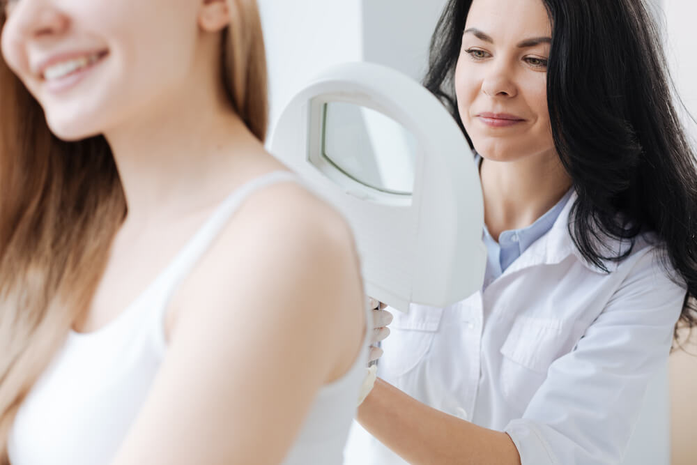 A dermatologist examines a patient&apos;s skin using a handheld diagnostic tool in a bright clinic setting. The patient, with long hair, appears relaxed while the dermatologist, dressed in a white lab coat, focuses on the examination. This image represents skin health assessments and the importance of professional skincare consultations.