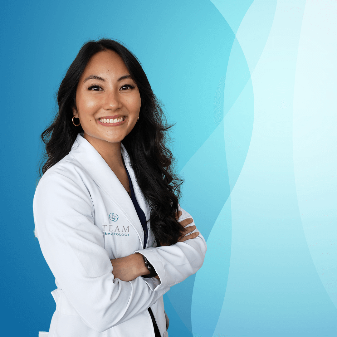 A smiling female dermatologist in a white lab coat stands confidently with her arms crossed. She has long, dark hair and is set against a soft blue gradient background. The image conveys professionalism and warmth, ideal for a dermatology practice.